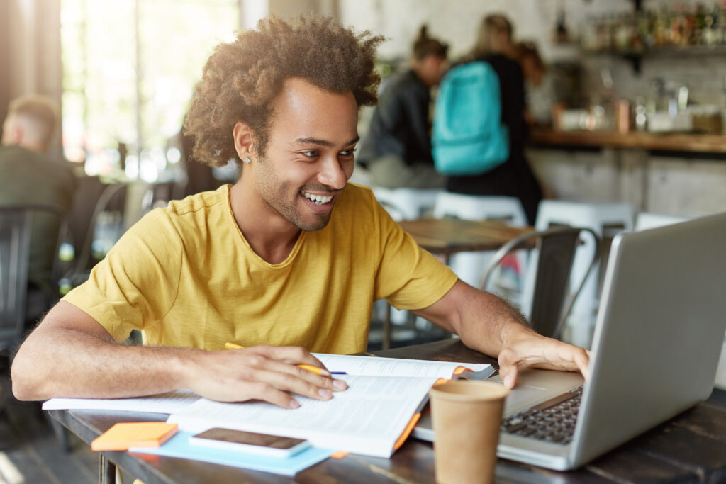 foto-interna-de-um-estudante-feliz-sexo-masculino-com-cabelo-encaracolado-vestido-casualmente-sentado-na-cafeteria-trabalhando-com-tecnologias-modernas-enquanto-estuda-olhando-com-um-sorriso-no-caderno-recebendo-